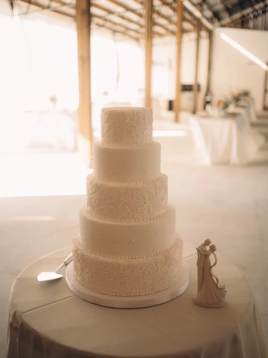 A five-tier white wedding cake with piped scrollwork on a draped table inside the barn