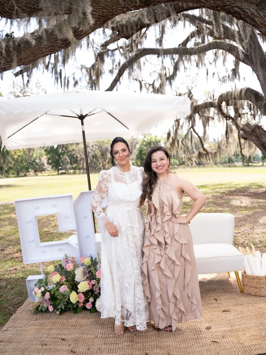 Two guests standing together under a moss-draped oak beside illuminated marquee letters and a floral arrangement