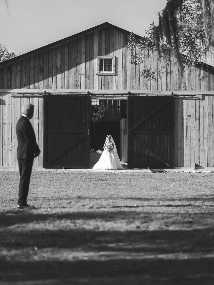 Black and white photograph of a bride holding her bouquet in the barn doorway as a guest watches from the lawn