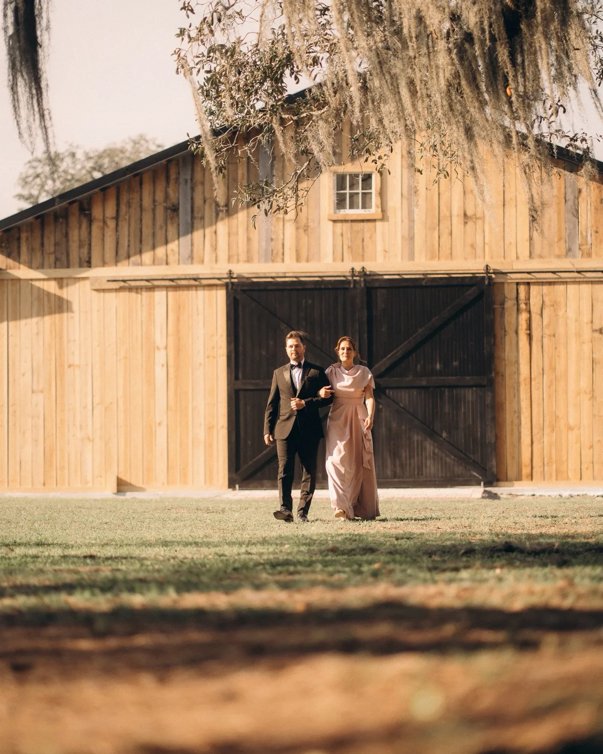 A couple in formal dress walking arm in arm from the barn's open doors across the lawn, Spanish moss hanging overhead